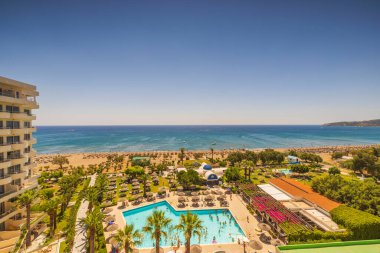 Açık havuzu, yeşil ağaçları ve bitkileri sonsuz mavi deniz su yüzeyinde bulutsuz mavi gökyüzü arka planıyla birleşen güzel panoramik otel manzarası. Rodos. Yunanistan. 07.15.2022.