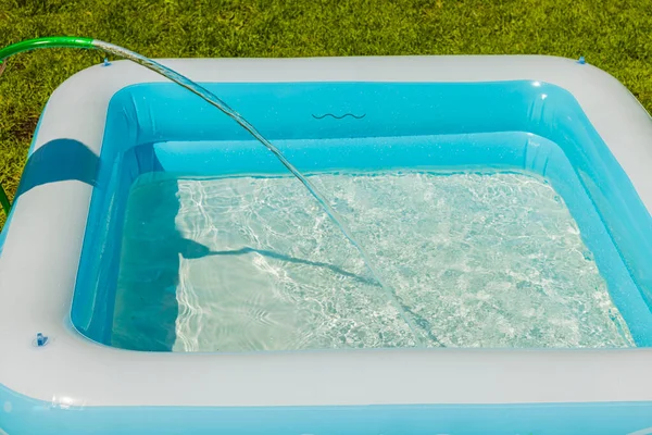 View of children\'s swimming pool being filled with water from garden hose for swimming on warm summer day.