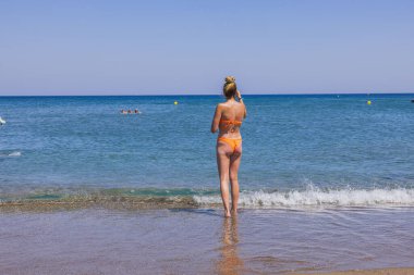 Kumsalda deniz suyuna gelen turuncu bikinili güzel bir kadın manzarası. Rodos. Yunanistan. 