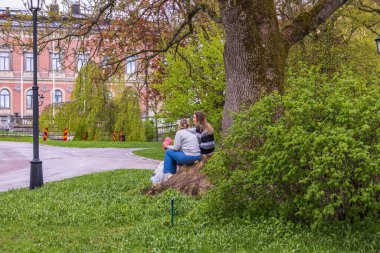 Beautiful view of two young woman sitting on roots of big old tree in park. Sweden. Uppsala. 05.14.2022.