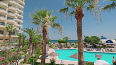 Beautiful view of hotel area with palm trees, vacationing tourists and  outdoor pool on Mediterranean coast. Rhodes. Greece. 08.06.2022.
