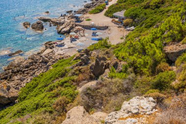 Beautiful view of people on private beach with sunbeds and umbrellas situated on rocky coastline. Greece. 