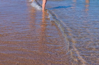 View of legs of man entering sea from shore of sandy beach. Greece.