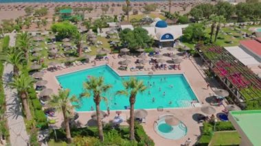 Beautiful view of hotel with tourists in around on sun beds under sun umbrellas at outdoor swimming pool and beach in Mediterranean sea. Rhodes. Greece. 07.28.2022
