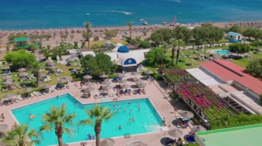 Beautiful top view of hotel with tourists in around on sun beds under sun umbrellas at outdoor swimming pool. Rhodes. Greece. 08.21.2022.