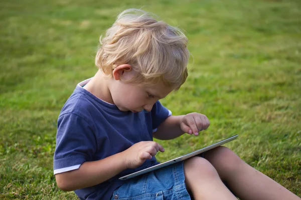 El chico rubio está sentado en el césped y jugando con la tableta de PC — Foto de Stock