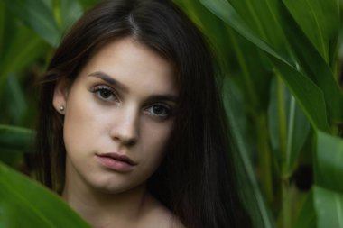 Attractive long haired tanned young woman is posing among the green plants closeup. Horizontally. 