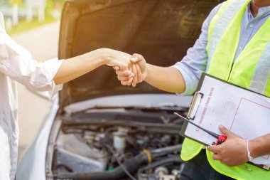Araba motoru arka planında güneş ışığı olan sigorta çalışanı ve müşterinin tebrik amaçlı el sıkışması..