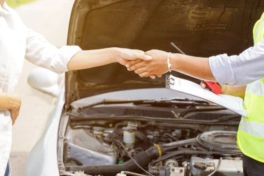 Araba motoru arka planında güneş ışığı olan sigorta çalışanı ve müşterinin tebrik amaçlı el sıkışması..