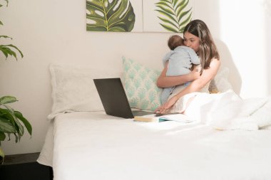 Latin anne dizüstü bilgisayar kullanırken bebeğine sarılıyor.