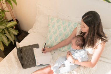 Latin anne bebeğinin yatağında dizüstü bilgisayar kullanıyor ve yazıyor.