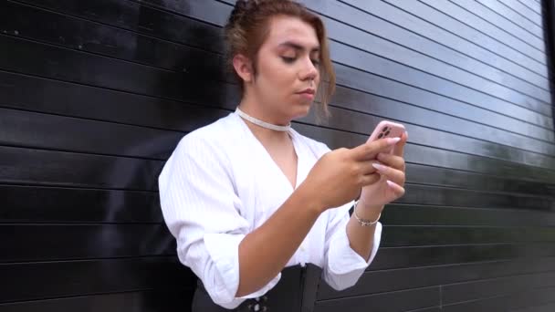 Небінарне використання телефону в її руках гоніння, спираючись на чорні ворота на вулиці — стокове відео