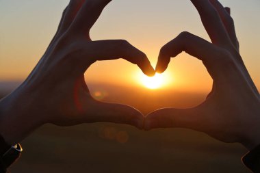 Günbatımı gökyüzünde, alacakaranlık vakti, güneş ışığı ellerden geçerken kalp şeklinde genç bir çocuğun elleri. Sevgi dolu kalp şeklinde eller. Aşk konsepti