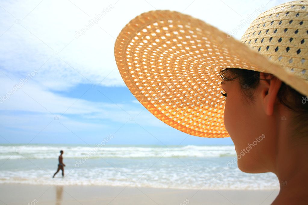 Beautiful woman with big straw hat looking at a man walking on the beach
