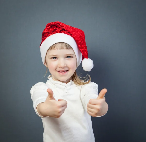 Başparmak gösterilen Noel Baba şapkalı küçük kız gülümseyerek — Stok fotoğraf