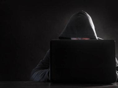 Kapüşonlu hacker karanlık arka planda dizüstü bilgisayarın arkasında oturuyor