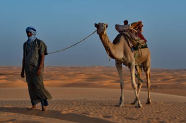 Shingetti. Moritanya 'da. Ekim 05, 2021. Ulusal elbiseli yalnız bir Mağripli Sahra Çölü 'nün kumlu kumullarında tek hörgüçlü bir deveye önderlik ediyor..
