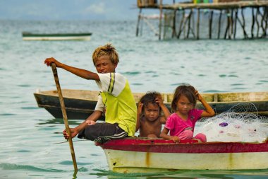 Semporna. Malezya. 30 Kasım 2018. Kürekli üç adam, deniz çingenelerinden oluşan bir balıkçı köyündeki kulübelerin arasında bir teknede yelken açıyorlar. Balıkçılar erken yaşlardan itibaren tekne yönetimi becerilerine sahipler..