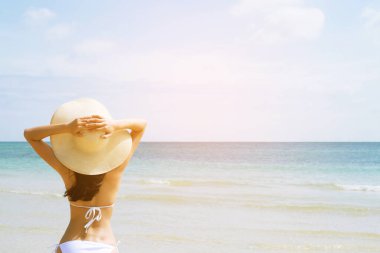 Güneş şapkası ve bikiniyle bronz tenli genç bir kadın plaj seyahatinin keyfini çıkarırken kollarını başının üstüne kaldırıyor. Ve sıcak yaz gününde deniz manzaralı hayatın tadını çıkar..