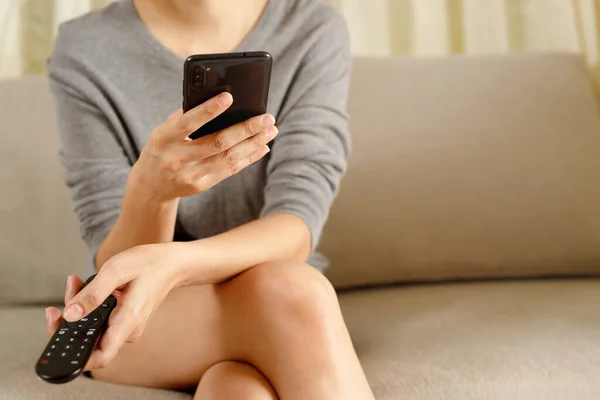 young woman using television remote control and mobile smart phone  is sitting on a sofa. on or off tv. Leave space to write a description of the message.