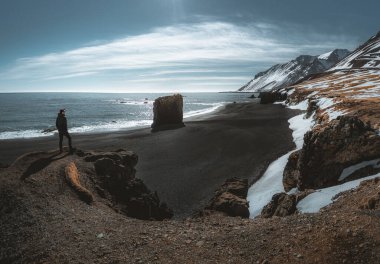 Person standing at black sand beach in Iceland. Fauskasandur near village of Djupivogur. Black sand beachwith snow and beautiful sunny weather. Seastack monolith.