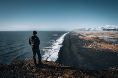 Person standing at Dyrholaey lighthouse in Iceland looking out over the black sand beach below during winter with snow and beautiful sunny weather.