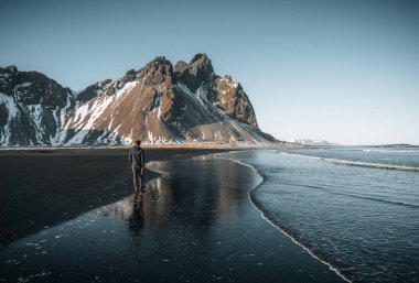 Gün batımında sahilde duran genç turist ve İzlanda 'daki Stoksnes Burnu' ndaki Vestrahorn Dağı 'nın muhteşem yansıması. Güzel kar kaplı dağlar. Konum: Stoksnes pelerini