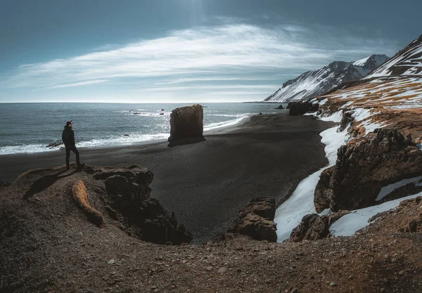 Personne debout à la plage de sable noir en Islande. Fauskasandur près du village de Djupivogur. Plage de sable noir avec neige et beau temps ensoleillé. Monolithe Seastack. — Photo