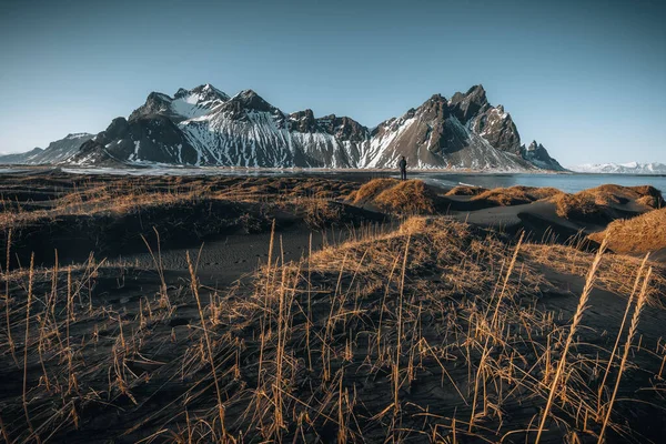 Ung man turist står på stranden under solnedgången runrise och vacker reflektion av Vestrahorn berg på Stokksnes udde på Island. Vackra snötäckta berg. Plats: Stokksnes udde — Stockfoto