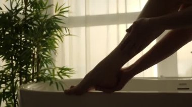 silhouette of female legs against the background of a large window with curtains. girl smears cream on her leg