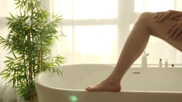 The girl smears her leg with cream in the bathroom against the backdrop of a large window with curtains — Stock videók
