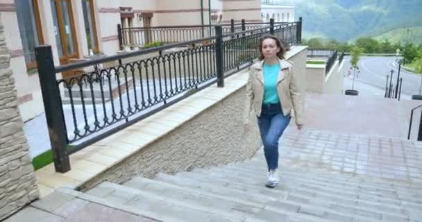 A woman in a white leather jacket and jeans climbs up the stairs on the street street — Video