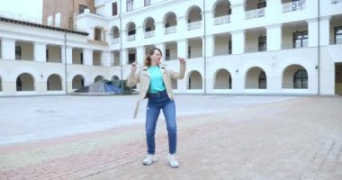 A young attractive woman in a white leather jacket and jeans dances and jumps happily in a suburban square