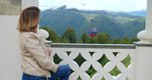 A woman in a white leather jacket is watching the movement of the funicular enjoying the scenery in a mountain resort — 비디오