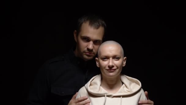 Eine glatzköpfige Frau und ihr Mann umarmen sich und blicken vor dunklem Hintergrund in die Kamera. Kopierraum — Stockvideo
