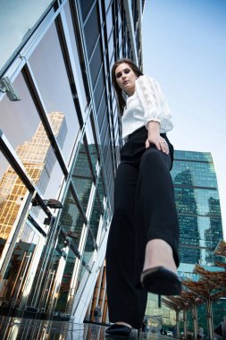 Sokaktaki genç iş kadını, arkasında cam ayna olan beyaz bluzlu ve siyah pantolonlu.