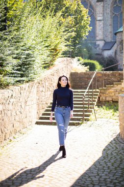 Siyah saçlı, arkasında ağaçlar olan beyaz bir kız kaldırım taşlarında yürüyor.