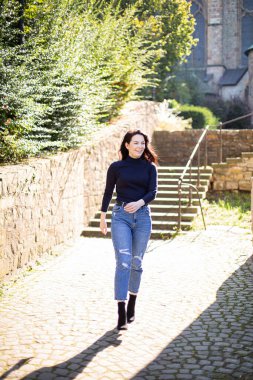 Siyah saçlı, arkasında ağaçlar olan beyaz bir kız kaldırım taşlarında yürüyor.