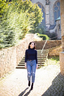 Siyah saçlı, arkasında ağaçlar olan beyaz bir kız kaldırım taşlarında yürüyor.