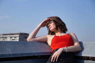 Koyu saçlı beyaz Avrupalı kadın çatıda oturuyor, arkasında açık gökyüzü var.