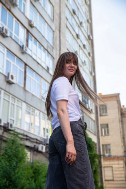 Arkasında binalar olan, siyah saçlı, gülümseyen beyaz kız.