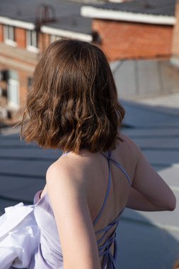 Esmer, kumral, çatıda oturan leylak elbiseli genç kadın.