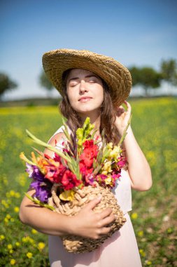 Kırsalda elinde çiçeklerle saman şapkalı, uzun saçlı, açık renk elbiseli beyaz bir kadının portresi.