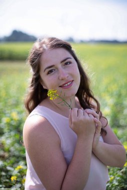 Kırsalda elinde çiçekle uzun saçlı, açık renk elbiseli beyaz bir kadının portresi.