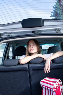 Koltukların arkasındaki açık araba bagajında koyu sarı, zayıf genç bir kadının portresi.