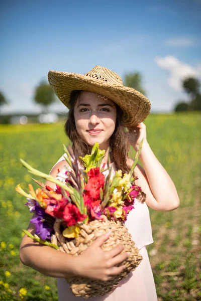 Kırsalda elinde çiçeklerle saman şapkalı, uzun saçlı, açık renk elbiseli beyaz bir kadının portresi.