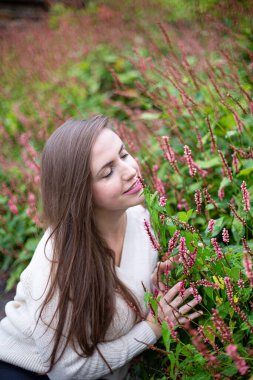 Beyaz Avrupalı kadın, çiçeklerin yanında, gözleri kapalı beyaz kazak giyiyor.