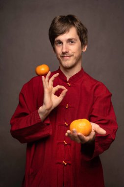 Genç yakışıklı, uzun boylu, zayıf, kahverengi saçlı, omuzlarında mandalina, elinde turuncu, gri arka planda kırmızı gömlekli adam.