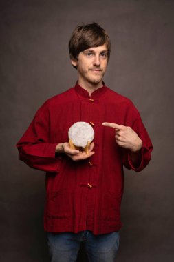 Genç, uzun boylu, zayıf, kahverengi saçlı, gri arka planda kırmızı gömlekli, pu erh çaylı pastayı işaret eden beyaz bir adam.