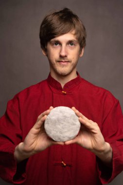 Genç, uzun boylu, zayıf, kahverengi saçlı, elinde pu erh çay kekiyle gri arka planda iki eli kırmızı gömlekli genç bir adam.